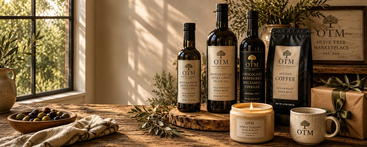 Olive Tree Marketplace olive oils, balsamic vinegar, artisan coffee, a lit candle, a bowl of olives and a wrapped gift arranged on a rustic wooden table in warm window light